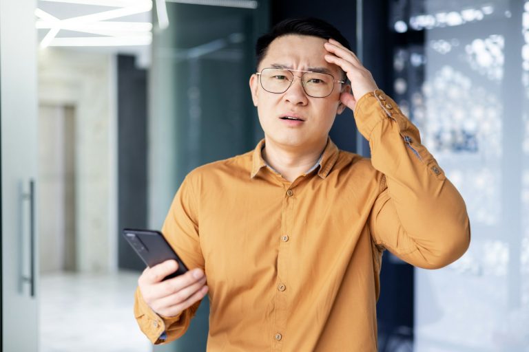 Unzufriedener asiatischer Mann mit Handy im Büro – MakeMore App Best Practices