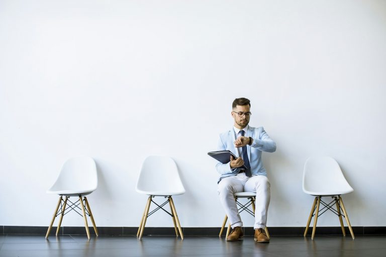 Young man sitting in the waiting room with a folder in hand and checking time before an interview